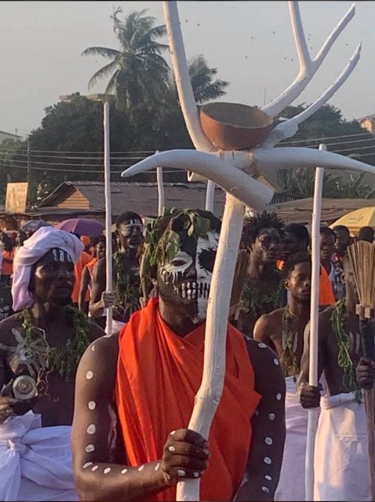 Man in orange with white body paint carrying a large white antler-shaped shrine object above the crowd during Fetu Afahye, Cape Coast