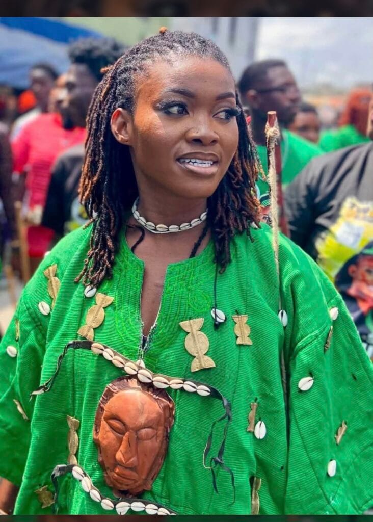 Young woman in a green cloth with a cowrie necklace and a small carved mask pendant, in the Fetu Afahye crowd, Cape Coast